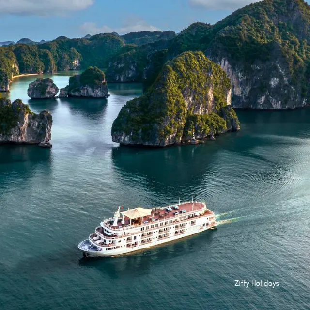 Aerial view of a large, white, multi-deck overnight cruise ship sailing through the dense network of dramatic limestone karst islands and emerald waters of Halong Bay, Vietnam.