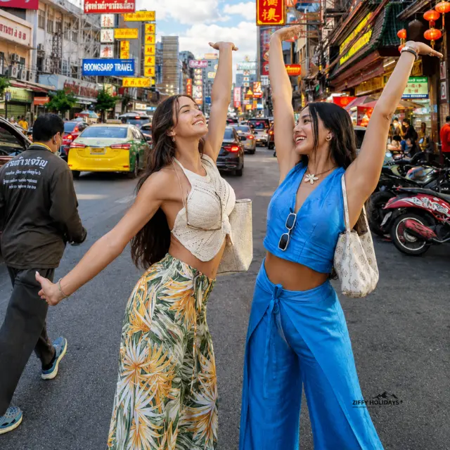 Two joyful women tourists posing with raised arms on the busy Yaowarat Road in Chinatown, Bangkok, Thailand.