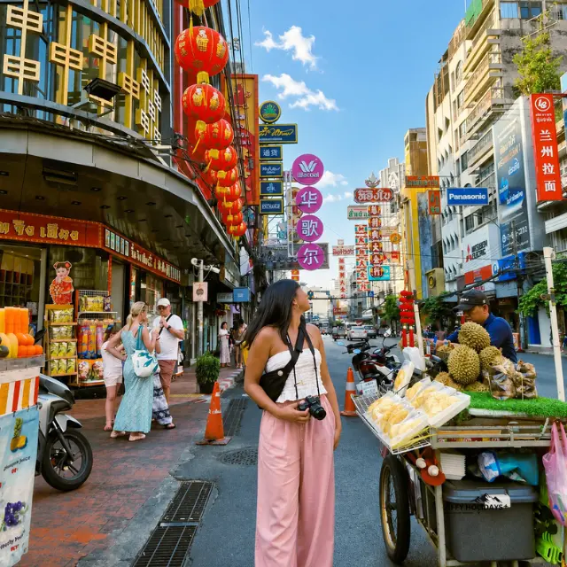 Female tourist looking at a street food cart selling fresh durian fruit under red lanterns in Chinatown, Bangkok.