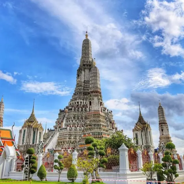 Detailed central spire of Wat Arun temple surrounded by lush green gardens under a cloudy blue sky in Bangkok, Thailand.