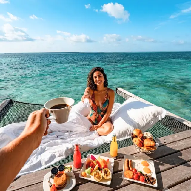 Happy woman sitting on an overwater hammock net enjoying breakfast and coffee over crystal clear ocean in Maldives.