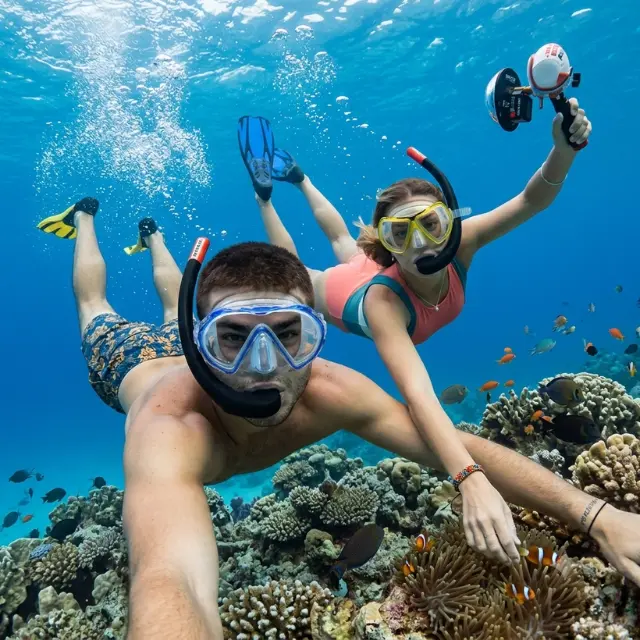 Couple wearing masks and flippers snorkeling underwater near vibrant coral reefs with tropical clownfish in Maldives.