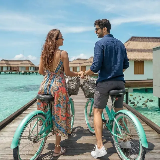 Romantic couple holding hands while riding turquoise bicycles on a wooden boardwalk leading to overwater bungalows in Maldives.
