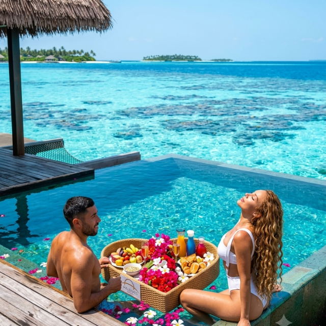 Couple relaxing in a private overwater infinity pool with a heart-shaped floating breakfast tray filled with fresh fruit and flowers in Maldives.