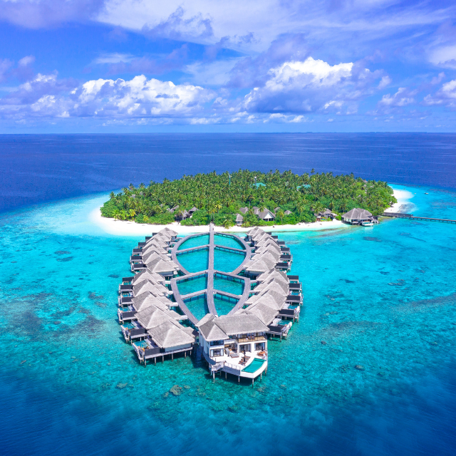 Aerial drone shot of a tropical green island surrounded by overwater villas and clear blue water in the Maldives.